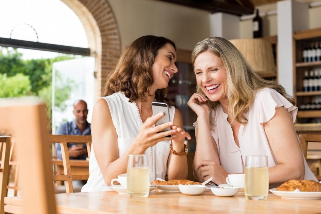 Female showing her friend something funny on her phone, sat with food and drinks